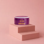 A purple cosmetic cream jar with gold lettering reading 'R.R.M WAH BEAUTY CREAM' sits on a two-tiered dusty pink pedestal against a matching pink background.